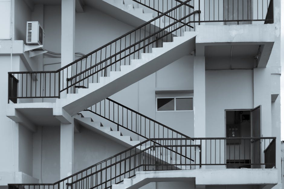 A multi-level external staircase of a modern white building featuring concrete steps and black metal railings, arranged in a zigzag pattern with landings visible at each floor level. The staircase spans vertically along the facade, with the lower section leading to a small door on the ground floor, and the upper sections extending upward behind the railings. A window is positioned between the stairs, and an air conditioning unit is mounted on the wall near the staircase. The environment appears to be an outdoor area, possibly part of a residential or commercial property, with natural or diffused lighting emphasizing the clean, minimalist architectural design. This scene may relate to on-site or private waste management activities, commonly involving the use of external access points for rubbish collection by specialist services such as Rubbish Removal Harrow, to facilitate efficient and accessible disposal procedures for multi-storey buildings.