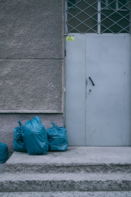A group of four blue plastic rubbish bags, tightly tied and filled with waste, are stacked against a textured grey concrete wall outside a building. The bags are placed on a concrete pavement in front of a light grey metal door that has a black handle and a small lock or latch underneath. To the upper right of the door is a rectangular window with a diamond-patterned metal grille. The scene appears to be part of a building's external service area, likely used for waste disposal or collection. The lighting is natural, suggesting daytime, and the environment is clean and orderly, consistent with private waste handling services such as those provided by Rubbish Removal Harrow, supporting alternative rubbish collection methods away from local authority services.