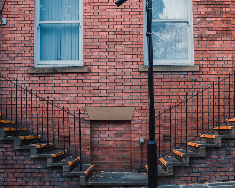 A multi-level external staircase of a modern white building featuring concrete steps and black metal railings, arranged in a zigzag pattern with landings visible at each floor level. The staircase spans vertically along the facade, with the lower section leading to a small door on the ground floor, and the upper sections extending upward behind the railings. A window is positioned between the stairs, and an air conditioning unit is mounted on the wall near the staircase. The environment appears to be an outdoor area, possibly part of a residential or commercial property, with natural or diffused lighting emphasizing the clean, minimalist architectural design. This scene may relate to on-site or private waste management activities, commonly involving the use of external access points for rubbish collection by specialist services such as Rubbish Removal Harrow, to facilitate efficient and accessible disposal procedures for multi-storey buildings.