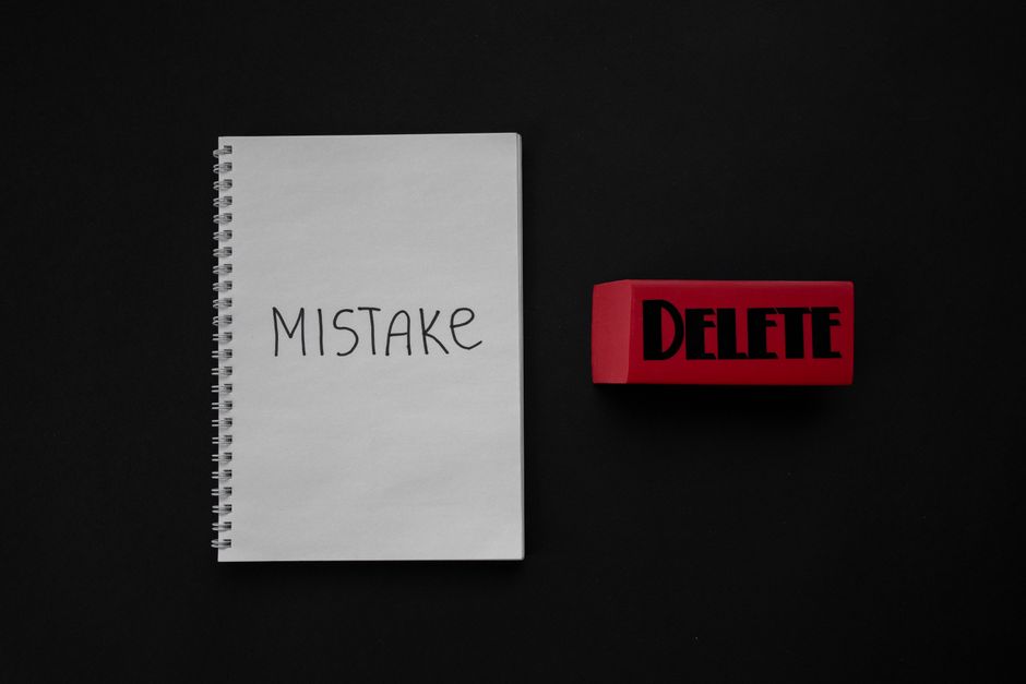 A white spiral-bound notepad placed on a black surface with the handwritten word 'MISTAKE' in large, black capital letters across the front. To the right of the notepad, there is a red rubber stamp with the word 'DELETE' printed in bold black letters. The overall scene has a professional and neutral tone, emphasizing the concept of mistake correction or removal, which is relevant to rubbish removal services such as those offered by Rubbish Removal Harrow in the context of managing unwanted waste or private disposal tasks. The lighting highlights the contrast between the black background and the white notepad, as well as the vivid red of the stamp, creating a clear and sharply focused visual suitable for an article discussing booking mistakes and cancellation fees for waste removal services, subtly supporting ideas related to alternative waste handling or on-site clearance options.