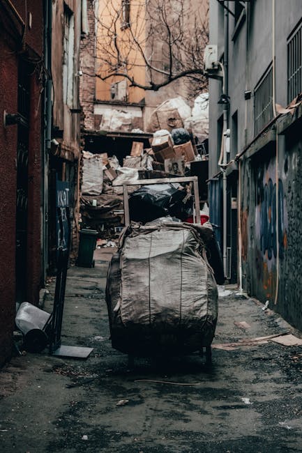 The image depicts a narrow urban alleyway cluttered with a large black garbage bag positioned in the foreground, on the uneven concrete ground that is stained and strewn with small debris. Behind the bag, there's a variety of discarded items and waste materials, including cardboard boxes, white plastic bags, and miscellaneous trash, piled up against a wooden and metal fence at the end of the alley. The surrounding environment features tall, aged buildings on either side, with visible graffiti on the right wall and an exposed utility box affixed to the front of the building on the right, indicating an area used for waste disposal or temporary rubbish storage. Bare tree branches extend overhead against a backdrop of a brown brick building, hinting at an urban setting in colder seasons. The overall scene is unkempt, emphasizing the need for waste collection services such as those provided by Rubbish Removal Harrow, which support private disposal and on-site clearance outside standard bin collection schedules in densely populated areas.