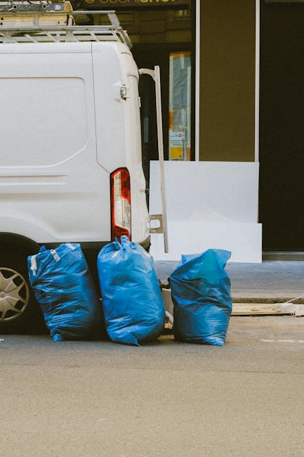 The image shows the rear side of a white delivery van parked on a paved street, with four large blue rubbish bags placed on the ground beside the vehicle's closed rear doors. The bags are made of plastic and appear to be filled with waste, exhibiting a crinkled and slightly shiny surface typical of compressed rubbish. The van is positioned close to the curb, and behind it, part of a building storefront with a glass door and window is visible, reflecting some interior light. To the right, a small white ramp or step is positioned leading up to the building's entrance. The environment suggests an urban setting, possibly related to private waste collection services like those offered by Rubbish Removal Harrow, which specializes in alternative rubbish disposal options outside official council services. The overall scene emphasizes on-site waste clearance, with the bags ready for removal by a private rubbish collection service, set against a neutral, everyday background of street and commercial premises.