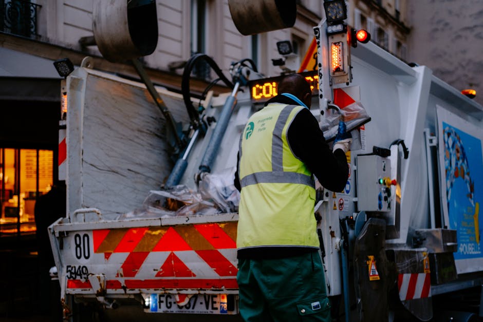 A worker wearing a high-visibility yellow vest with reflective stripes and blue gloves is operating a waste collection vehicle on an urban street during dusk or early evening. The worker's back is facing the camera as they handle controls on the side of the truck, which is loaded with a pile of mixed waste materials, including paper, plastic, and possibly fabric, visible in the open rear compartment. The waste appears loosely piled and contains various textures and colors, such as white, brown, and transparent plastics along with crumpled paper and fabric. The collection vehicle is predominantly white with orange and red reflective safety markings on its rear and sides, with some mechanical components and hydraulic parts visible near the worker. The background shows parts of urban infrastructure, including illuminated windows of a building and possibly street lighting, creating a dimly lit atmosphere. The scene depicts a routine private or municipal rubbish collection, aligning with waste removal services like those offered by Rubbish Removal Harrow, illustrating the process of on-site waste collection that supports alternative rubbish disposal methods outside of council bulk waste collection rules.