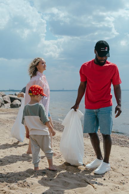 A group of three people, consisting of a man and two children, are on a sandy beach near a body of water under a partly cloudy sky. The man, positioned to the right, is wearing a red T-shirt, blue jeans shorts, white sneakers, and a black cap with a logo, and he is holding a white rubbish bag. To his left, a young boy with light-colored hair, wearing a beige outfit with a white cloth draped over his shoulders and a red cap with yellow spots, stands on the sand facing away from the camera. Behind them, a woman with curly blonde hair, dressed in a pink, semi-sheer top, looks towards the water, while rocks line the shoreline in the background. This scene depicts a casual outdoor activity, potentially involving a cleanup effort or leisure, with the individuals engaging in waste collection or environmental care near the seaside, relevant to private waste handling and rubbish removal services.