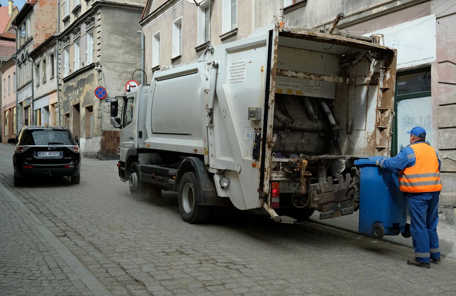 A white refuse collection truck is parked on a narrow cobblestone street alongside old multi-storey buildings with light-colored facades and traditional architectural details. The rear section of the truck, which appears rusted and used, is open and empty, designed for collecting waste. A worker dressed in blue coveralls and a high-visibility orange vest is positioned on the right side of the image, actively pushing a blue wheeled bin toward the truck's intake chute. The street features parked cars, including a black hatchback on the left, and has a slight incline. On the wall of the building directly behind the truck, there are traffic regulation signs, including a no-parking sign and a restricted access sign. The scene is lit with natural daylight, creating soft shadows and providing clear visibility of textures, such as the rough cobblestones, the weathered surface of the truck, and the worker's safety gear. This image illustrates an example of private waste collection and rubbish removal services in an urban setting, possibly as part of an on-site clearance or alternative waste handling managed by Rubbish Removal Harrow, emphasizing the practical process of rubbish collection outside of municipal services.