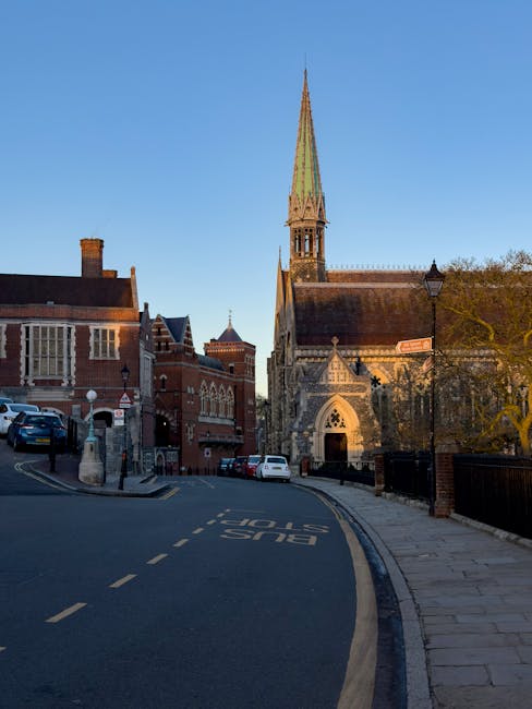 A narrow street in Harrow on the Hill with a sloping asphalt road marked 'BUS STOP' in white letters. On the left side, there are parked cars, a red brick building with white window frames, and a sidewalk with stone paving. On the right side, there is a church with a tall, pointed greenish spire, arched windows, and stone masonry, partially obscured by a large tree with budding leaves. A vintage-style streetlamp is situated near the church, and the scene is illuminated by natural daylight, with a clear blue sky overhead. The streetscape suggests an area with historic architecture, and Rubbish Removal Harrow could provide private waste disposal services for the local community, which often involves on-site clearance or independent rubbish collection to manage waste efficiently in such settings.