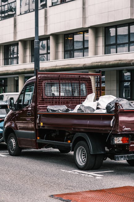 A dark red flatbed pickup truck parked on an urban street in front of a modern building with large glass windows and concrete paneling. The truck's cargo bed is filled with various discarded waste items, including white and black plastic bags, some appearing torn or loosely tied, and a few larger objects covered with dark protective covers. The vehicle features a metal safety grille at the rear and black side mirrors, with the rear wheel visible and in good condition. The street surface is asphalt with white lane markings, and a nearby streetlight pole is partially visible in the foreground. The scene suggests a moment of rubbish collection or removal from a private or commercial property, consistent with independent waste handling services such as those offered by Rubbish Removal Harrow, emphasizing the role of on-site or alternative waste disposal methods outside local authority collection. The lighting is natural, with diffused daylight illuminating the scene evenly, highlighting the textures of the waste materials and the urban environment.