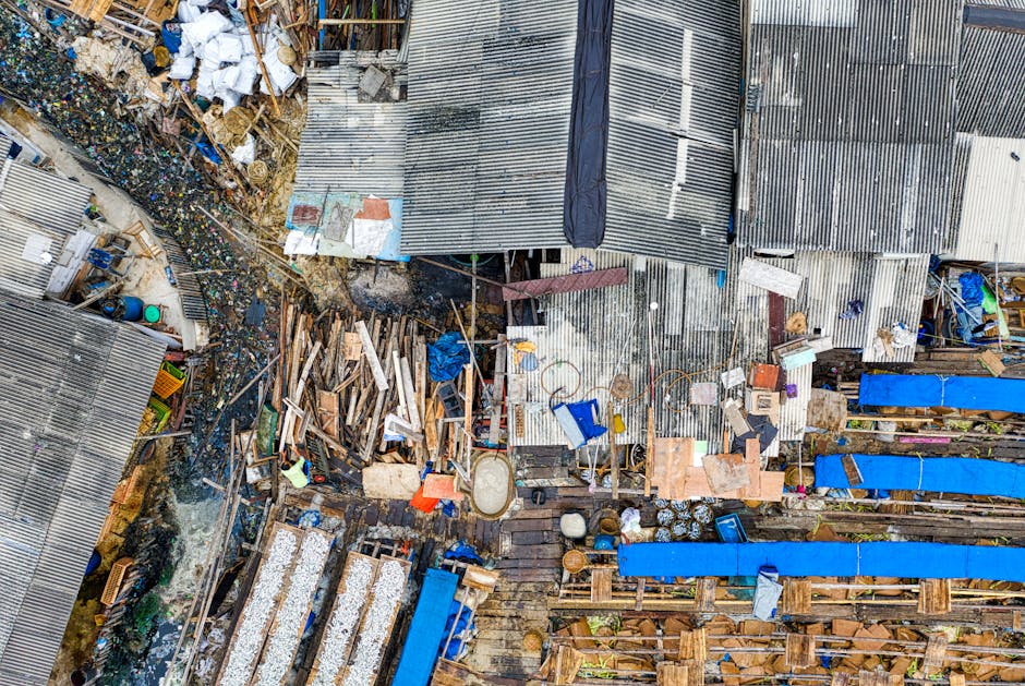 An aerial view of a cluttered yard adjacent to makeshift structures with corrugated metal roofs, showcasing an assortment of discarded materials including wooden planks, plastic buckets, and various piles of rubbish. The scene features uneven surfaces with areas of water or mud, and a mix of debris scattered across the space, reflecting a typical site for private waste collection or on-site clearance. The roofs are weathered, showing signs of rust and wear, with some sections covered by tarpaulins or plastic sheets. Several blue tarpaulins are stretched over parts of the wooden fencing, which appears hastily assembled. The environment indicates a busy, operational area where waste is being accumulated and managed, consistent with the context of independent rubbish removal services. The lighting is natural, likely overcast, adding a muted tone to the scene, which highlights the diversity of materials and textures present. This visual documentation aligns with the activities of companies like Rubbish Removal Harrow involved in alternative waste handling and rubbish clearance, providing a clear representation of a typical waste collection site in an informal setting.