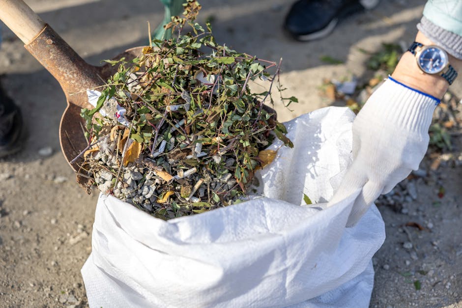 A person wearing a white glove and a wristwatch is holding a large white bag filled with garden debris, including small branches, twigs, and dry leaves. The bag appears to be made of durable woven material suitable for waste collection. The debris inside has various textures, with some leafy green stems and rough, twiggy fragments visible at the top. The person is standing outdoors on a gravel surface, with a metal shovel or spade resting in the bag, indicating active garden or yard waste removal. In the background, there are hints of other garden debris scattered on the ground, and a pair of dark shoes or boots is partially visible. This scene reflects a typical waste clearance operation, likely performed by a professional rubbish removal service such as Rubbish Removal Harrow, who provide alternative waste handling options for garden and yard waste disposal, especially for properties requiring private or non-council services.