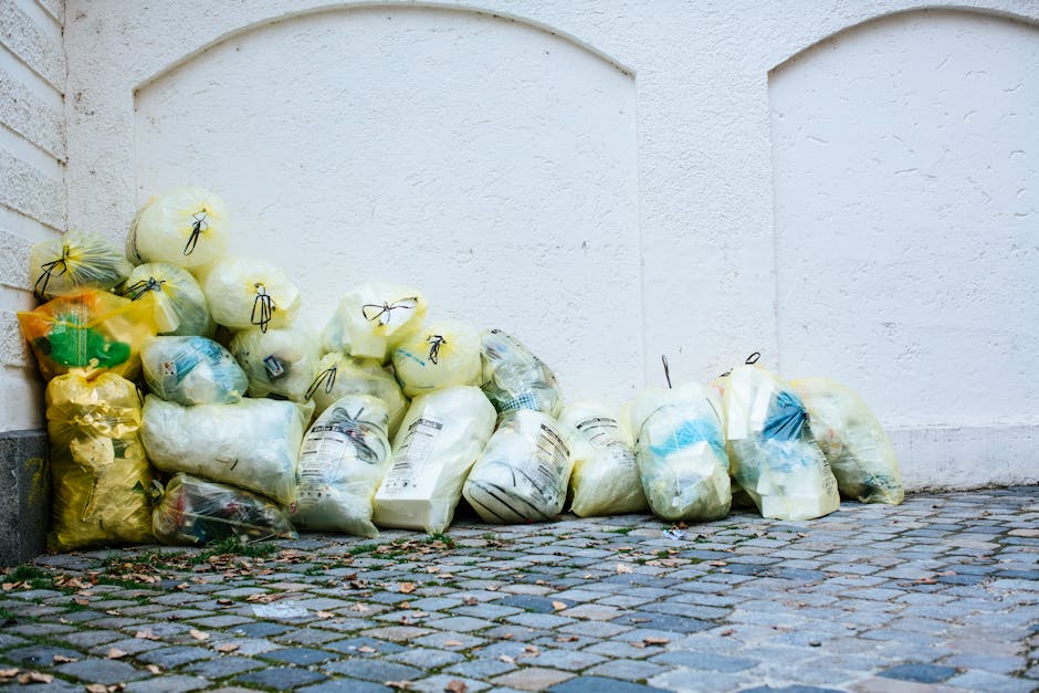 A white waste collection truck parked on a city street during daytime, loaded with various rubbish and debris. The truck's side panel displays the number 610 and the label 'OBEREAL'. The load includes black garbage bags, cardboard boxes, plastic containers, and miscellaneous waste materials piled towards the top of the truck. In front of the vehicle, on the pavement, there are additional black trash bags and loose debris. The background features a multi-storey brick building with several windows, some with metal grills, and a row of trees casting shadows on the facade. The scene indicates private waste collection or rubbish removal activities typical within urban environments, with the truck representing a service that handles rubbish disposal away from local authority services, aligning with independent waste clearance practices.